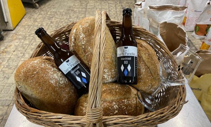 Twee flesjes Zwarte Parochie bier in een broodmand met vier broden en een leeg bierglas erin